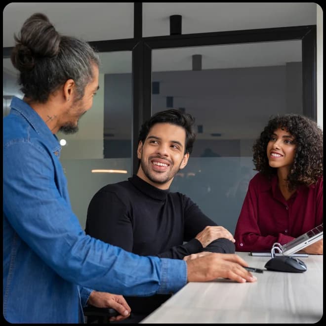 Three colleagues smiling and talking at a table in a bright office.