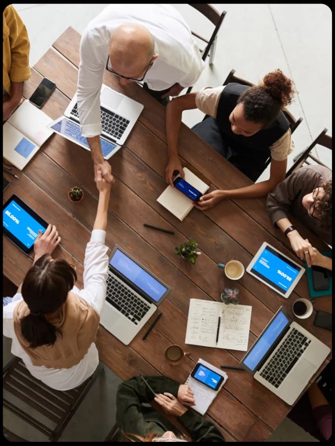 Overhead view of a team around a long table with laptops and tablets, two people shaking hands across it.