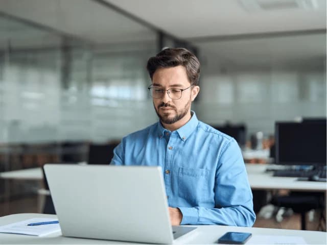 A man in a blue shirt and glasses working on a laptop at an office desk