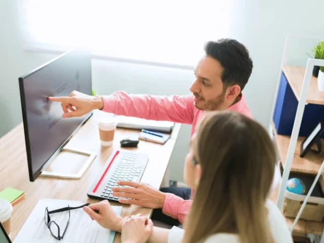 A man pointing at a desktop monitor while a woman beside him looks on