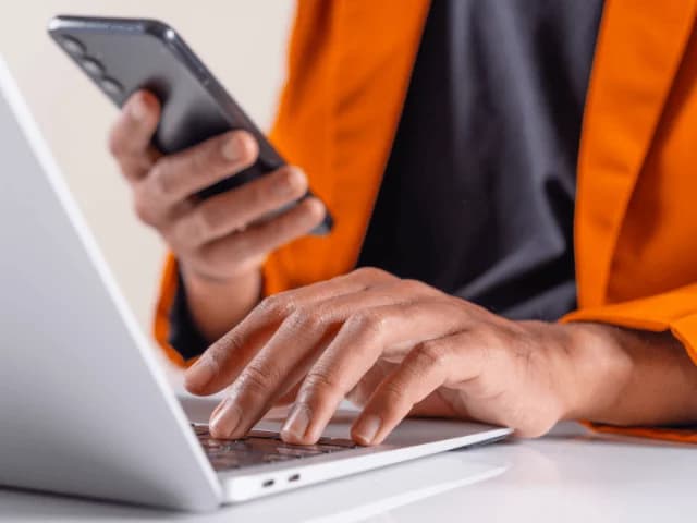 Hands typing on a laptop while one holds a phone