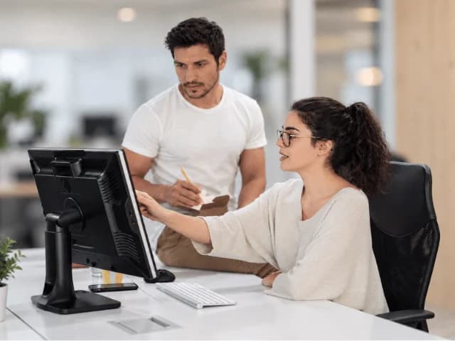 A man taking notes while a woman points at a desktop monitor in an open office
