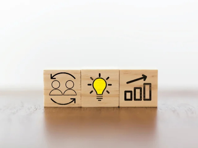 Three wooden blocks showing icons of two people, a light bulb and a rising bar chart