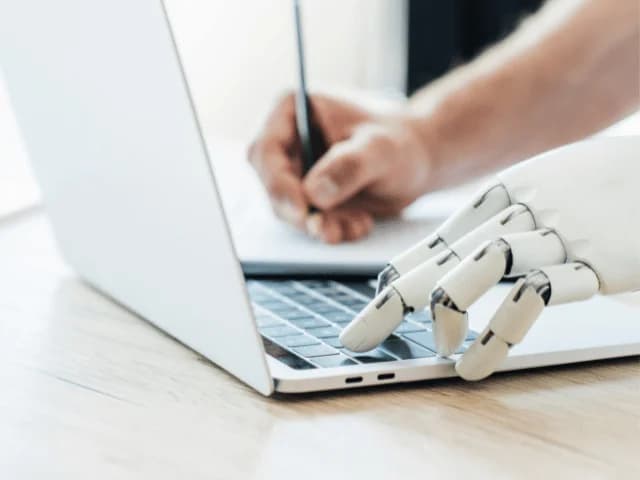 A hand writing with a pen next to a robotic hand typing on a laptop