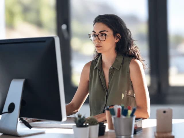 A woman with glasses working at a desktop computer in an office